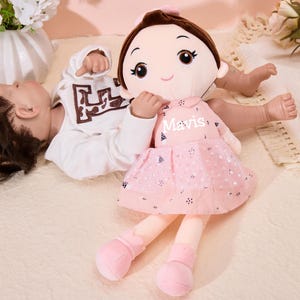 May include: A pink plush doll with a pink dress and black polka dots. The doll has brown hair, brown eyes, and a pink bow. The doll's name is "Mavis" and is lying next to a baby doll.