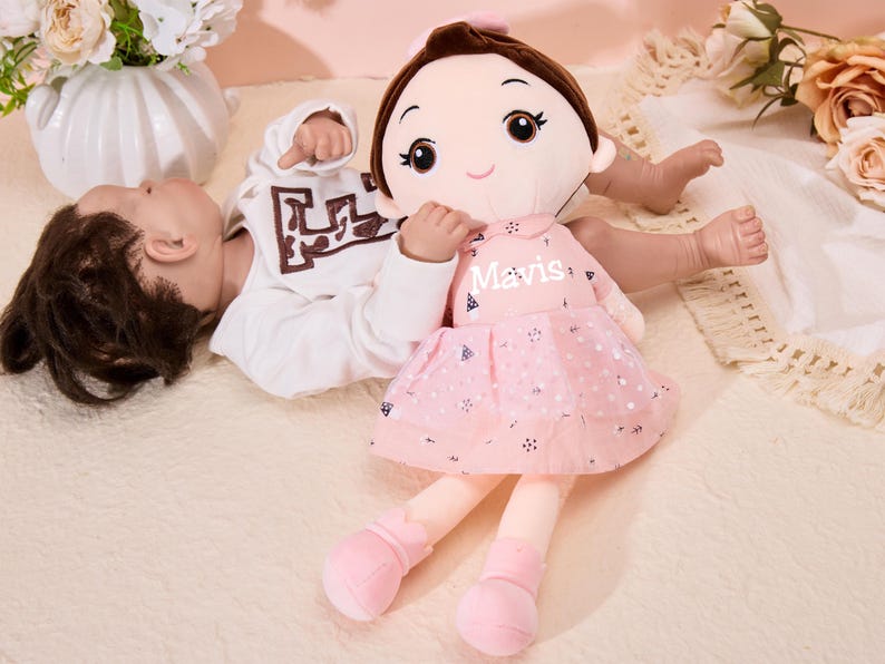 May include: A pink plush doll with a pink dress and black polka dots. The doll has brown hair, brown eyes, and a pink bow. The doll's name is "Mavis" and is lying next to a baby doll.
