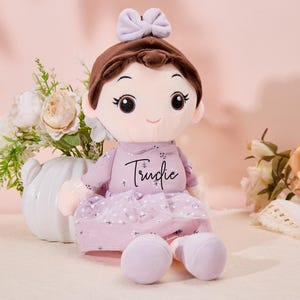 May include: A plush doll with brown hair, a purple dress, and a bow. The doll has a sweet expression and the name "Trudie" embroidered on the dress.