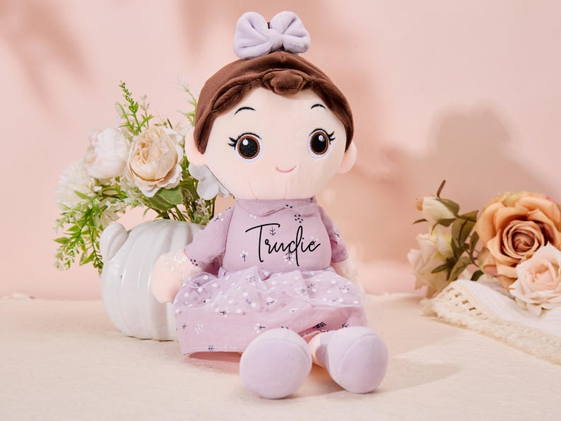 May include: A plush doll with brown hair, a purple dress, and a bow. The doll has a sweet expression and the name "Trudie" embroidered on the dress.