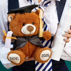 Puede incluir: Un oso de peluche marrón con birrete y toga de graduación, sosteniendo un diploma con una cinta amarilla. El oso tiene el nombre "RACHEL" y el año "2026" impresos en sus patas. El fondo es una persona con una camisa blanca y una corbata a rayas azules y blancas.