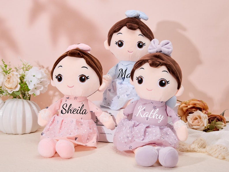 May include: Three plush dolls with brown hair and embroidered names. The dolls are wearing pink, purple, and blue dresses with floral patterns. The dolls are sitting on a white surface.