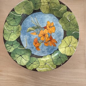 May include: A round ceramic plate featuring a central blue field decorated with a cluster of orange nasturtium flowers and stems. The border is adorned with a repeating pattern of large, rounded green leaves with visible veins on a dark background.