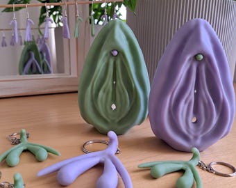 May include: A collection of handmade art pieces in shades of green and purple. The image features two large vulva sculptures, earrings, and keychains. The sculptures are on a wooden surface, with a plant in the background. The earrings and keychains are in the shape of a clitoris.