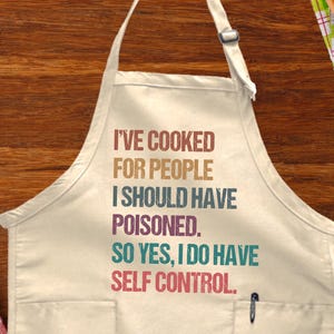 May include: Cream-colored apron with the text "I'VE COOKED FOR PEOPLE I SHOULD HAVE POISONED. SO YES, I DO HAVE SELF CONTROL." A pen is in the pocket. The apron is on a wooden surface with cooking utensils and food.