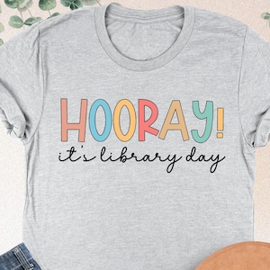 May include: A light gray t-shirt with the words "HOORAY! it's library day" in colorful letters. The shirt is paired with blue jeans and a brown hat. The words "ATHLETIC HEATHER" are printed at the top.