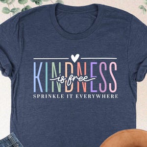 May include: Heather navy blue t-shirt with the words "KINDNESS is free, sprinkle it everywhere" in colorful text. The image also includes a pair of blue jeans and a brown hat.