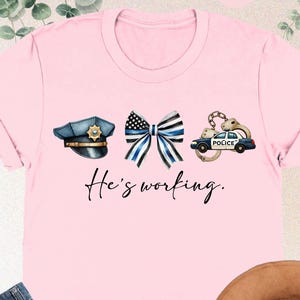 May include: Pink t-shirt with a blue police officer's hat, a blue and white ribbon, handcuffs, and a police car with the text "He's working."