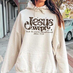 May include: Sand-colored hoodie with the text "Jesus wept but He never complained John 11:35" in a decorative font. The hoodie has a front pocket and a drawstring hood. The image also includes the word "SAND".