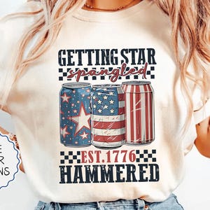 May include: Ivory-colored t-shirt with a patriotic design featuring three beverage cans decorated with stars and stripes. The text reads "GETTING STAR SPANGLED EST. 1776 HAMMERED". Additional text on the shirt says "MORE COLOR OPTIONS".