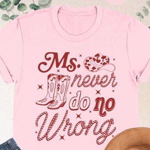 May include: A pink t-shirt with the text "Ms. never do no Wrong" in a western-style font, along with illustrations of cowboy boots and a hat. The image also includes a pair of blue jeans and a brown cowboy hat.