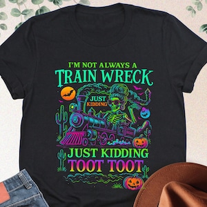 May include: Black t-shirt with a colourful Halloween graphic. The design includes a train, skeleton, pumpkins, and the text "I'm Not Always A Train Wreck Just Kidding Toot Toot". A pair of blue jeans and a brown hat are also in the image.