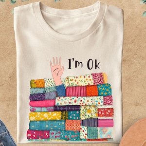 May include: Cream-colored t-shirt with the text "I'm Ok" and a graphic of a hand emerging from a stack of colorful fabric. The fabric has various patterns and colors, including blue, pink, and yellow.