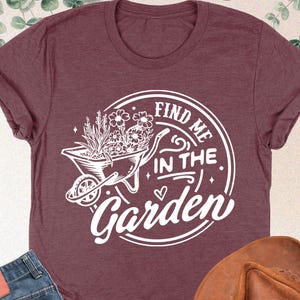 Puede incluir: Una camiseta de color marr&oacute;n rojizo con un gr&aacute;fico blanco de una carretilla llena de flores y el texto "Find me in the Garden".