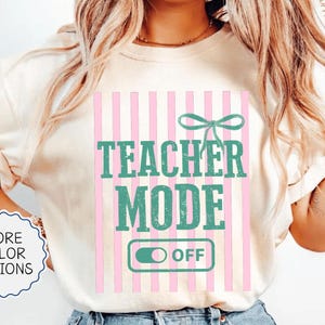 May include: Ivory-colored t-shirt with the text "TEACHER MODE OFF" in teal and pink. The design includes a pink and white striped background and a teal bow. The shirt also has a "MORE COLOR OPTIONS" button.
