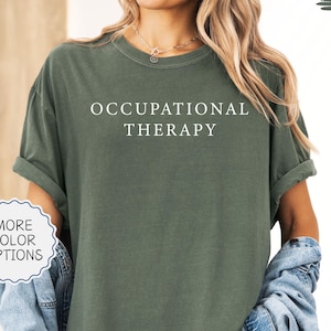 May include: Moss green t-shirt with the words "OCCUPATIONAL THERAPY" in white. The shirt has rolled-up sleeves and is paired with a denim jacket. A button with "MORE COLOR OPTIONS" is visible. The shirt's color is described as moss.