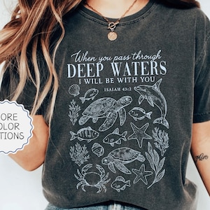 May include: Pepper-colored t-shirt with a white graphic featuring marine life and the text "When you pass through deep waters, I will be with you, Isaiah 43:2." The shirt has a crew neck and short sleeves. "More color options" is also displayed.