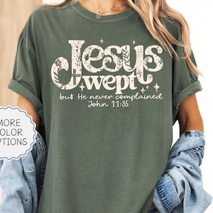 May include: Moss green t-shirt with the text "Jesus wept but He never complained John 11:35" in a decorative font. The shirt has rolled-up sleeves and a relaxed fit. The words "More Color Options" are in a white circle.