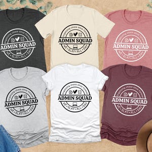 May include: A collection of t-shirts in various colors, including heather grey, white, and mauve. Each shirt features a circular graphic with the text "ADMIN SQUAD" and the phrase "organizing chaos since forever, we slay all day."