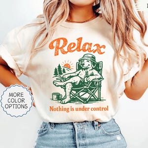 May include: Ivory-colored t-shirt with the word "Relax" in orange above a green illustration of a dog relaxing in a camping chair. The text "Nothing is under control" is below the illustration. The shirt has a crew neck.