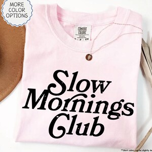 May include: A pale pink t-shirt with the words "Slow Mornings Club" printed in black. The shirt has a crew neck and short sleeves. A delicate necklace with a small pendant rests on the shirt. The words "More Color Options" are in a circle on the top left.