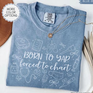 May include: A blue jean colored t-shirt with the phrase "BORN TO YAP forced to chart" in white. The shirt features medical-themed illustrations. A gold necklace with a pendant is draped across the shirt.