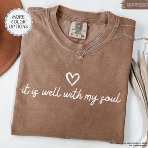 May include: Espresso-colored t-shirt with the phrase "it is well with my soul" in white script, accompanied by a heart symbol. The shirt has a crew neck and short sleeves. A silver necklace with a pendant is draped on the shirt.