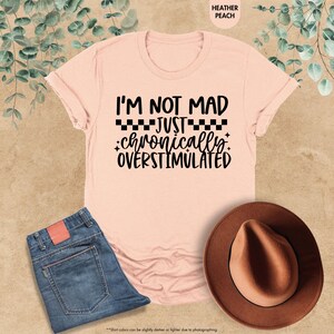Op de afbeelding: Een perzikkleurig t-shirt met de tekst "I'M NOT MAD JUST chronically OVERSTIMULATED" in het zwart. Het shirt wordt getoond met een blauwe spijkerbroek en een bruine hoed. De kleur van het shirt wordt omschreven als heather peach.