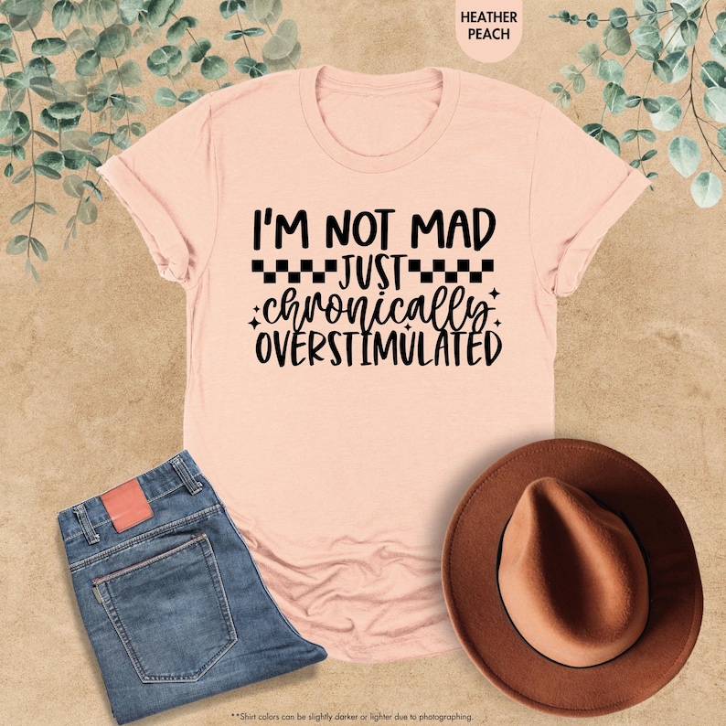 Op de afbeelding: Een perzikkleurig t-shirt met de tekst "I'M NOT MAD JUST chronically OVERSTIMULATED" in het zwart. Het shirt wordt getoond met een blauwe spijkerbroek en een bruine hoed. De kleur van het shirt wordt omschreven als heather peach.