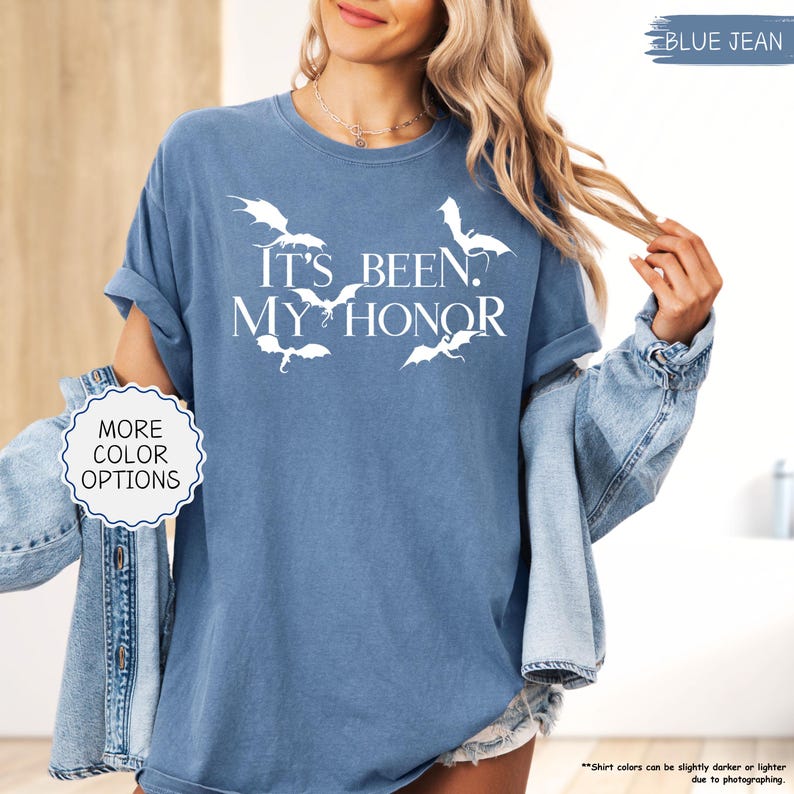 Pu&ograve; includere: Una maglietta blu jeans con la frase bianca "IT'S BEEN. MY HONOR", insieme a grafiche di draghi bianchi. La maglietta ha le maniche arrotolate e fa parte di una collezione di abbigliamento. L'immagine include anche un pulsante "MORE COLOR OPTIONS".