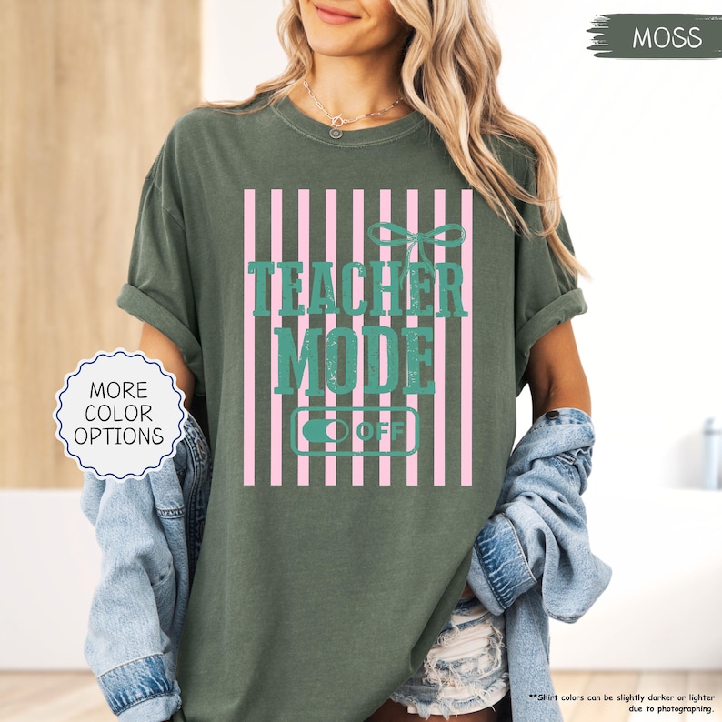 May include: Moss green t-shirt with the text "TEACHER MODE OFF" in teal, set against a pink striped background. The shirt has rolled-up sleeves and a relaxed fit. "MORE COLOR OPTIONS" is in a white circle.