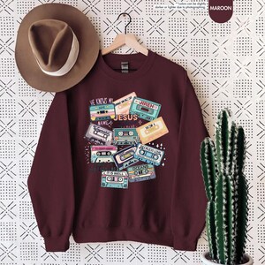 May include: Maroon sweatshirt with a graphic of vintage cassette tapes with Christian song titles printed on them. The titles include "He Knows My Name", "Waymaker", "Jesus", "Jireh", "So Am I", "Reckless Love", "It Is Well", and "Oceans".