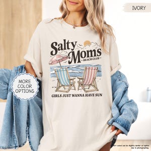 May include: Ivory-colored t-shirt with the text "Salty Moms - Beach Club" and the phrase "Girls Just Wanna Have Sun." The graphic features beach chairs, an umbrella, and a beach scene. The shirt is a relaxed fit.