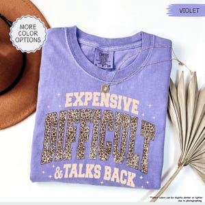 May include: A violet Comfort Colors t-shirt with the text "EXPENSIVE, DIFFICULT, & TALKS BACK" in a tan and leopard print design. A gold necklace and a brown hat are also visible.