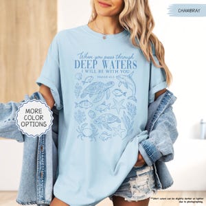 May include: Light blue t-shirt with the text "When you pass through deep waters, I will be with you" and Isaiah 43:2. The shirt features a blue illustration of sea creatures, including turtles, fish, and a dolphin. The shirt is a chambray color.