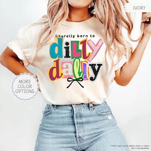 K&ouml;nnte beinhalten: Elfenbeinfarbenes T-Shirt mit dem farbenfrohen und verspielten Schriftzug "literally born to dilly dally". Das Design beinhaltet eine schwarze Schleife und die W&ouml;rter sind in verschiedenen Farben. Das Shirt hat einen Rundhalsausschnitt und kurze &Auml;rmel.