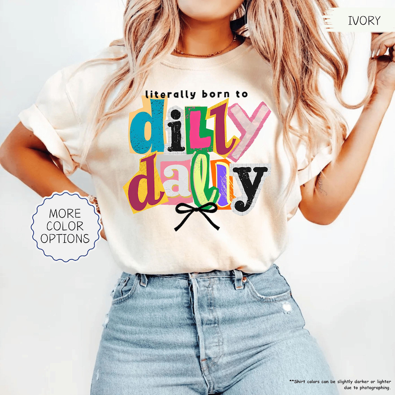 K&ouml;nnte beinhalten: Elfenbeinfarbenes T-Shirt mit dem farbenfrohen und verspielten Schriftzug "literally born to dilly dally". Das Design beinhaltet eine schwarze Schleife und die W&ouml;rter sind in verschiedenen Farben. Das Shirt hat einen Rundhalsausschnitt und kurze &Auml;rmel.
