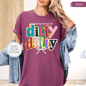 K&ouml;nnte beinhalten: Beerenfarbenes T-Shirt mit dem Schriftzug "literally born to dilly dally" in einem farbenfrohen, geschichteten Design. Das Shirt hat hochgekrempelte &Auml;rmel und wird mit einer Jeansjacke getragen. Ein Knopf auf der linken Seite zeigt "MORE COLOR OPTIONS".