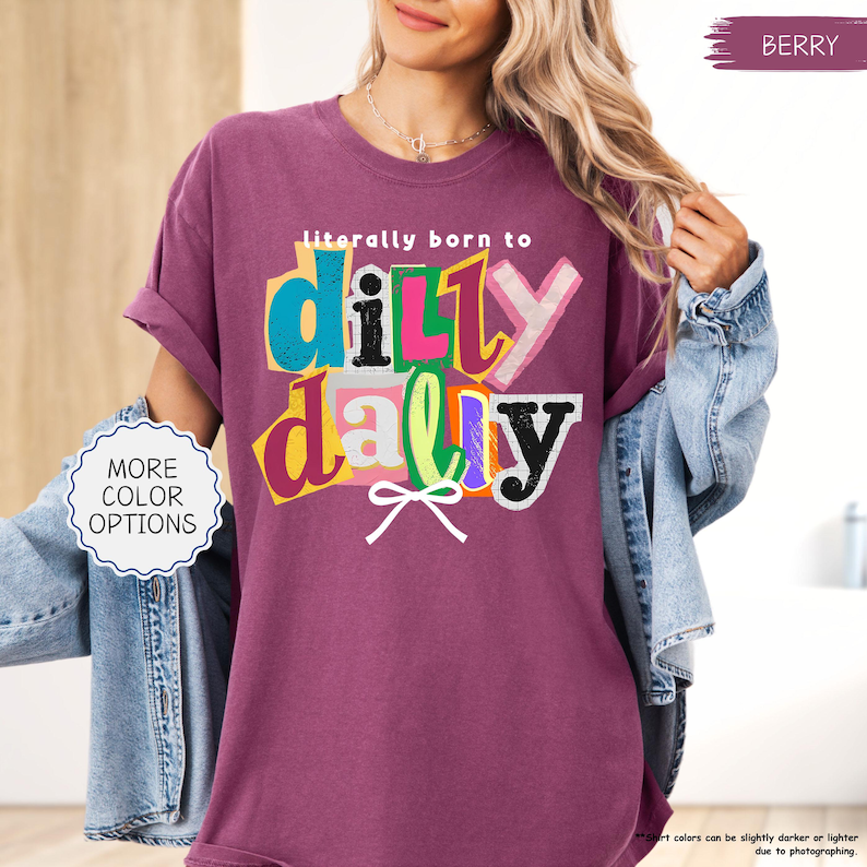 K&ouml;nnte beinhalten: Beerenfarbenes T-Shirt mit dem Schriftzug "literally born to dilly dally" in einem farbenfrohen, geschichteten Design. Das Shirt hat hochgekrempelte &Auml;rmel und wird mit einer Jeansjacke getragen. Ein Knopf auf der linken Seite zeigt "MORE COLOR OPTIONS".