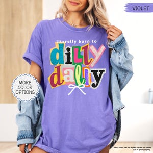 K&ouml;nnte beinhalten: Ein violettes T-Shirt mit dem Schriftzug "literally born to dilly dally" in einem farbenfrohen, ausgeschnittenen Stil. Das Shirt hat kurze &Auml;rmel und eine lockere Passform. &Uuml;ber dem Shirt wird eine Jeansjacke getragen. Der Text "MORE COLOR OPTIONS" ist ebenfalls sichtbar.
