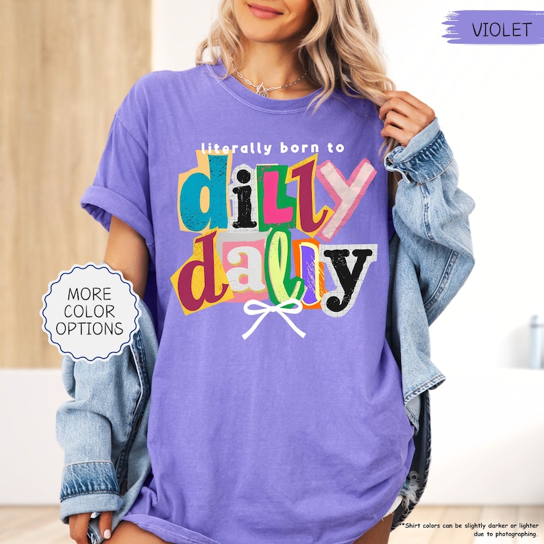 K&ouml;nnte beinhalten: Ein violettes T-Shirt mit dem Schriftzug "literally born to dilly dally" in einem farbenfrohen, ausgeschnittenen Stil. Das Shirt hat kurze &Auml;rmel und eine lockere Passform. &Uuml;ber dem Shirt wird eine Jeansjacke getragen. Der Text "MORE COLOR OPTIONS" ist ebenfalls sichtbar.