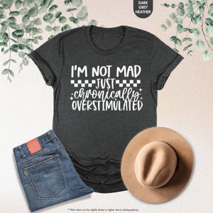 Op de afbeelding: Donkergrijs heather T-shirt met de witte tekst "I'M NOT MAD JUST chronically OVERSTIMULATED". De afbeelding bevat ook een spijkerbroek en een bruine hoed.