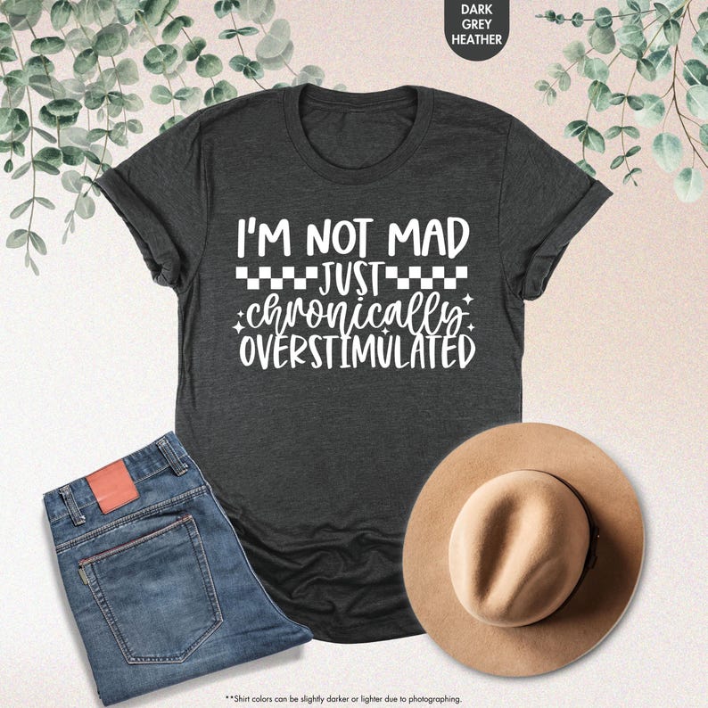 Op de afbeelding: Donkergrijs heather T-shirt met de witte tekst "I'M NOT MAD JUST chronically OVERSTIMULATED". De afbeelding bevat ook een spijkerbroek en een bruine hoed.