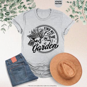 Puede incluir: Una camiseta gris con un gr&aacute;fico negro de una carretilla llena de flores y el texto "Find me in the Garden".