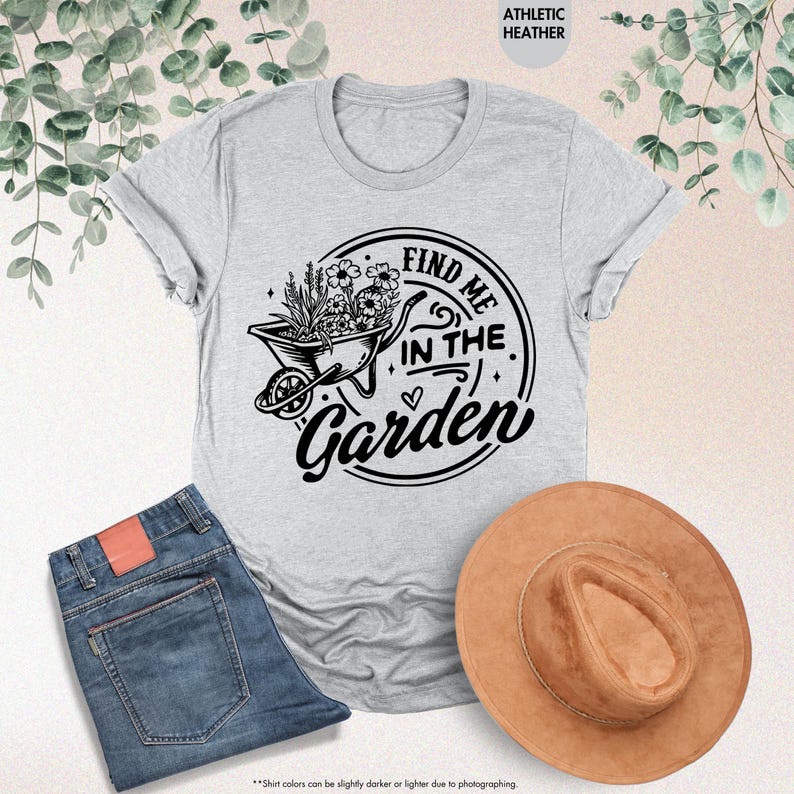 Puede incluir: Una camiseta gris con un gr&aacute;fico negro de una carretilla llena de flores y el texto "Find me in the Garden".