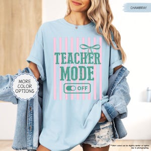 May include: Light blue t-shirt with the text "TEACHER MODE OFF" in green, set against pink vertical stripes. A green bow is above the text. The shirt is a chambray color and has "MORE COLOR OPTIONS" text on the left.