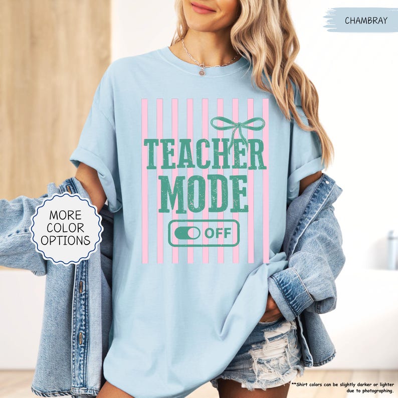 May include: Light blue t-shirt with the text "TEACHER MODE OFF" in green, set against pink vertical stripes. A green bow is above the text. The shirt is a chambray color and has "MORE COLOR OPTIONS" text on the left.