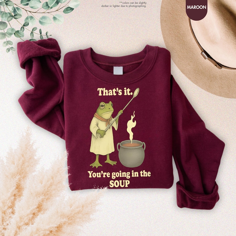 Pu&ograve; includere: Felpa bordeaux con un ranocchio dei cartoni animati che tiene un cucchiaio sopra una pentola di zuppa. Il testo "That's it. You're going in the SOUP" &egrave; stampato sul davanti. Maniche lunghe e girocollo.