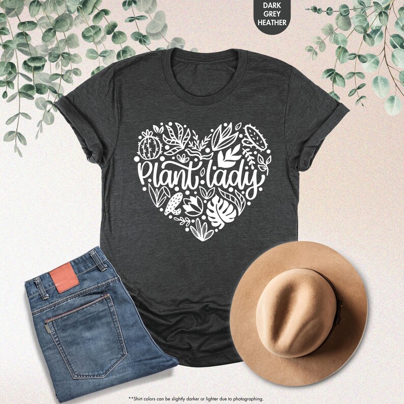 May include: A dark heather grey t-shirt with a white heart-shaped design featuring the text "Plant lady" and various plant illustrations.