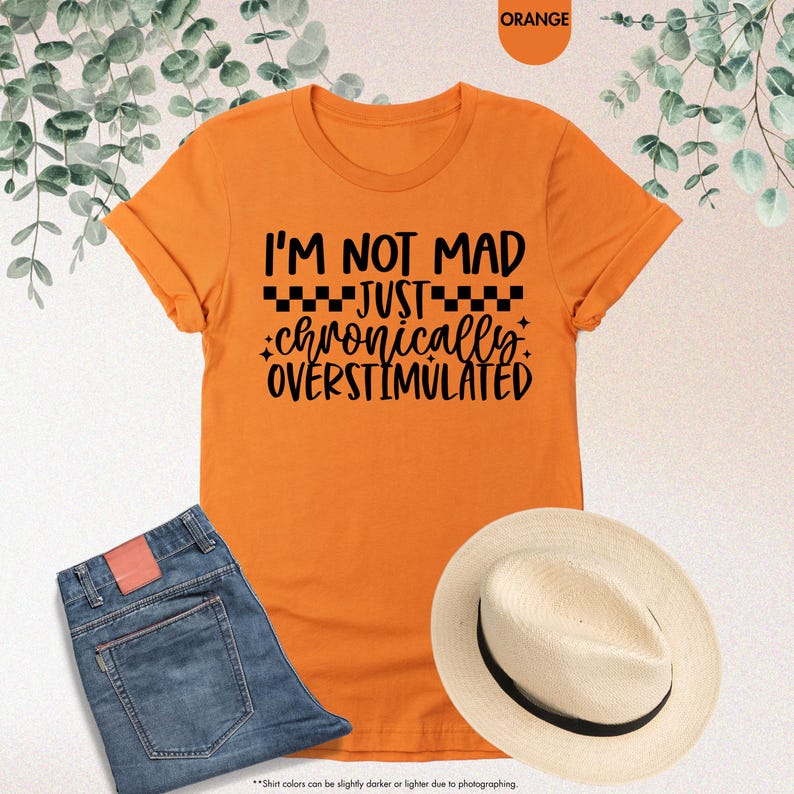 Op de afbeelding: Oranje T-shirt met de zwarte tekst: "I'M NOT MAD JUST chronically OVERSTIMULATED". De afbeelding toont ook een blauwe spijkerbroek en een strohoed. Het T-shirt is oranje.